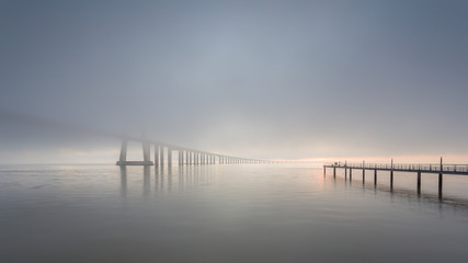 Obraz premium Vasco da Gama Bridge and pier over Tagus River in Lisbon, Portugal, at sunrise with a dense fog.