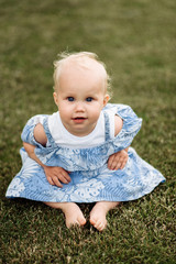 A very cute little blue-eyed baby girl in a blue dress sits on the grass barefoot and smiling