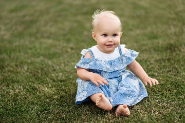 A very cute little blue-eyed baby girl in a blue dress sits on the grass barefoot and smiling, on the nature