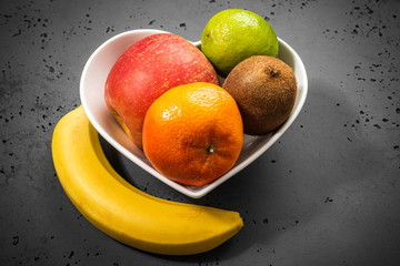 Heart-shaped bowl with fruit on stone background. The concept of healthy eating, eating fruit. Bowl with apple, mandarin banana. A bowl filled with fruit. Fruit and vitamins for the heart.