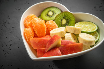 Heart-shaped bowl with fruit on stone background. The concept of healthy eating, eating fruit. Bowl with apple, mandarin banana. A bowl filled with fruit. Fruit and vitamins for the heart.