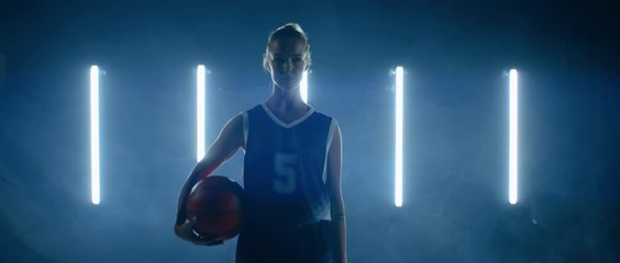 Caucasian female basketball player posing in generic uniform, holding ball in her hands against bright lights. 4K UHD 60 FPS SLOW MOTION shot on ARRI Alexa Mini with Atlas Orion 2x Anamorphic lens