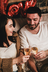 Young smiling happy couple in love in comfortable home clothing clinking glasses of champagne and celebrating holiday at home. Valentines day celebration concept