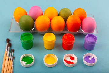 Colorful handmade Easter eggs and jars of multicolored gouache with tassels.. Top view of an open transparent box. Isolated on a blue background. Preparing for Easter.