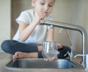 Portrait of little caucasian girl gaining a glass of tap clean water. Kitchen faucet. Kid pouring...