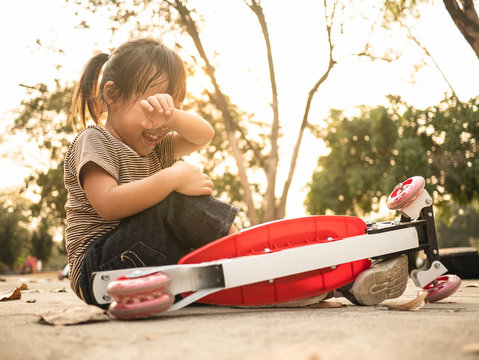 Cute Asian little girl sitting on the ground after falling off her scooter at summer park. Healthy outdoor sport for young child.