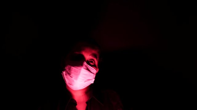 Woman in the surgical medical mask in the darkness and red light. Personal forced home quarantine isolation from Coronavirus or covid 19 concept. Apocalypses and depression mood of global epidemic.