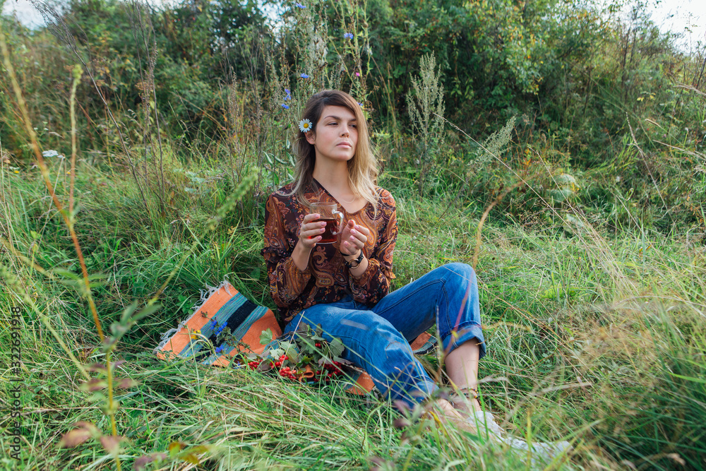 Wall mural Beautiful woman with camomile flower in hair sitting on the grass and drinking tea outdoors