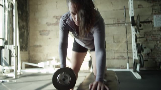 Camera follows weight as woman does dumbell rows in industrial-style gym