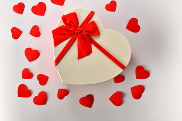 Box. Heart-shaped gift box with red ribbon stands in the middle on a white background with little red cardboard hearts. The view from the top. Valentine's day and Birthday
