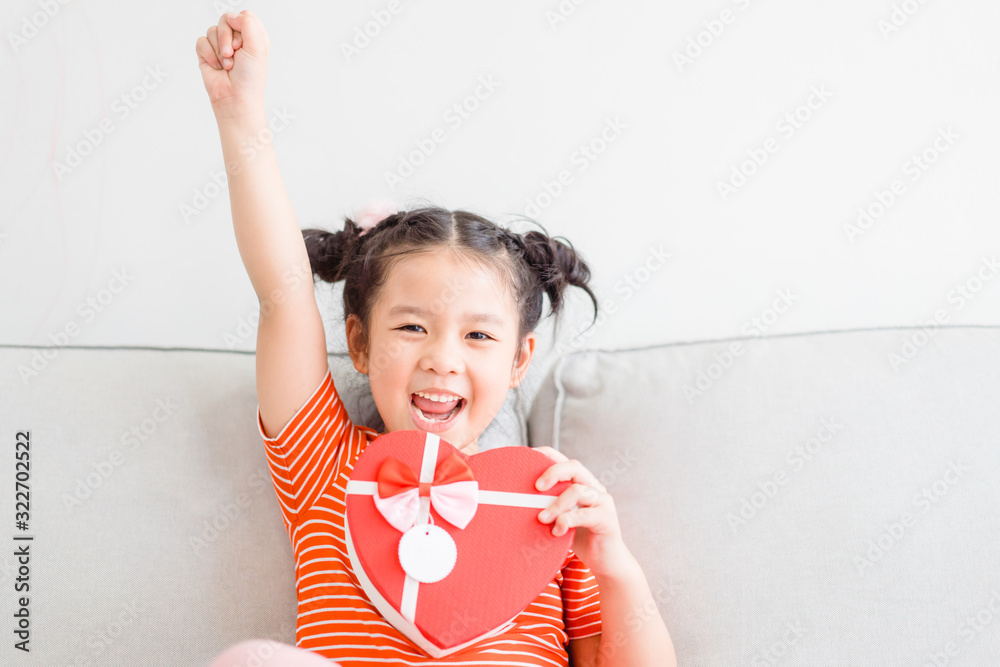 Wall mural Little asian girl raised hands smile and excited and holding red gift box heart shape in sofa at home on white wall background.Child girl holding gift box in Valentine, Christmas and New year concept.