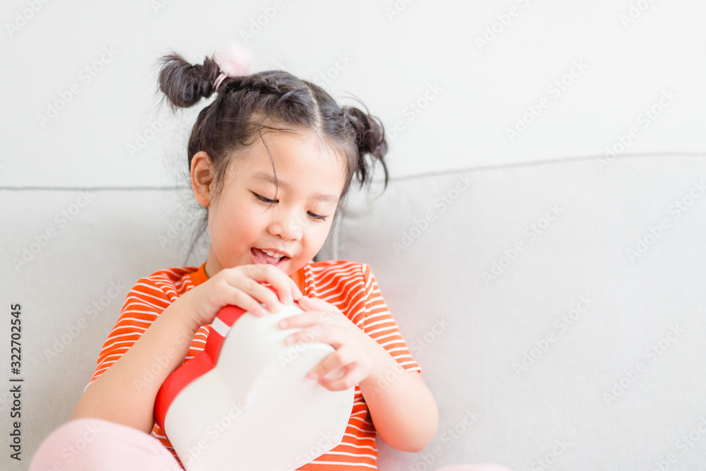 Wall mural Little asian girl Happy smiley excited face and open red gift box in heart shape in sofa at home on white wall background.Child girl holding gift box in Valentine day, Christmas and New year concept.