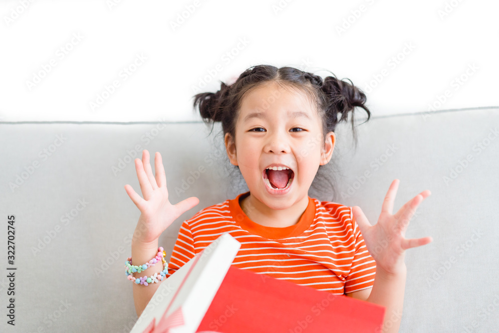 Wall mural Little asian girl smile and excited and holding red gift box on sofa in living room background.child holding gift box in Christmas and New year concept.