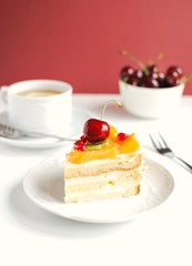 Slice of cake with fruits and cherry on top. Yoghurt cake with fruits served on white plate with cup of tea, bowl with cherries.