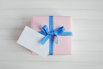 Light pink gift box with blue ribbon on white wooden background. Present with blank tag for message. Close-up top view.