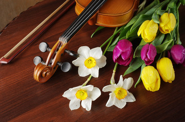 Violin with a bow, a bouquet of daffodils and tulips on a wooden table.