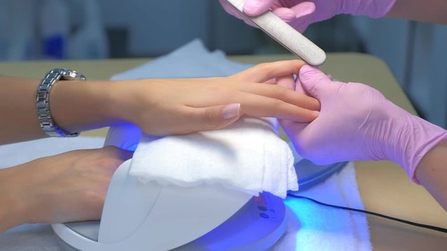 Manicurist master in gloves polishing client's nails using file, hands closeup. Professional manicure in beauty salon with gel polish covering. Drying shellac in UV lamp. Beauty industry concept.