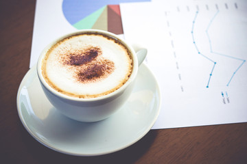 A white coffee cup with a creamy foam placed on the desk with graph data documents. Office work concepts