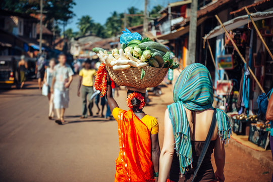 An Indian woman in red Sari carries a large basket of vegetables on her head. Tourists and locals in India. A crowded street in Gokarna, Karnataka.