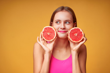 Beauty portrait of a lovely young woman with long hair standing over yellow background, showing slices of a grapefruit.