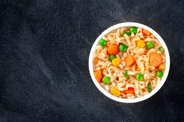 Ramen cup, instant soba noodles in a plastic cup, with green peas and carrot, overhead shot with copy space on a black background