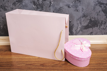 pink paper bag and heart shaped gift box lying on wooden floor against concrete wall background....