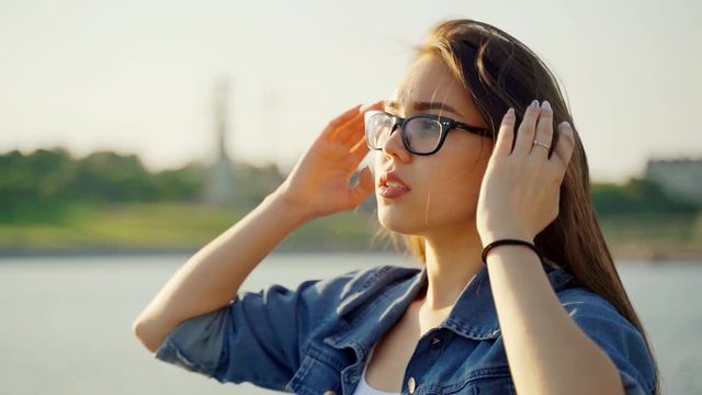 A girl with glasses and a denim jacket walks along the bay and straightens her hair. The wind blows dark hair shining in the sun at sunset. Macro video. Hair in the wind. Slow motion