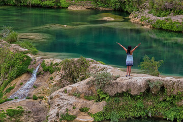 Rear of a woman standing over the rock stretching her hands and enjoying the waterfall, waterfall...