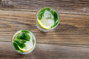 Two glasses of lemonade with lemon, mint and ice cubes on wooden background. Summer drink