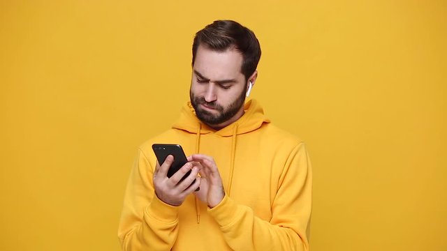 Handsome bearded young guy 20s in streetwear hoodie isolated on yellow background studio. People sincere emotions lifestyle concept. Listen music in modern earphones enjoy dance hold in hand cellphone