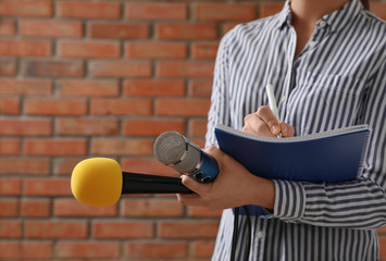 Journalist with microphones and notebook near brick wall, closeup