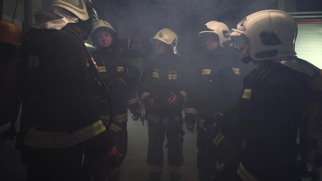 group of five firefighters wearing full equipment. relaxed chatting after work well done. smoke and fire trucks in the background. Name tag translation is fireman and model's last name.
