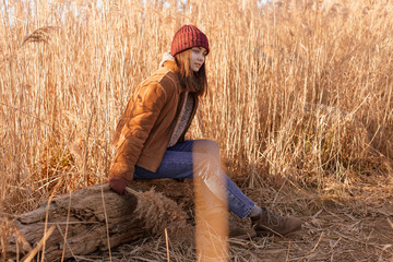 Naklejka na ściany i meble Thoughtful lady sits on a log and looks away. Young woman in field brown high grass.