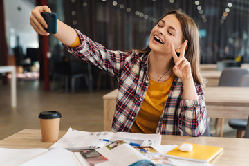 Image of joyful girl taking selfie on cellphone and gesturing peace sign