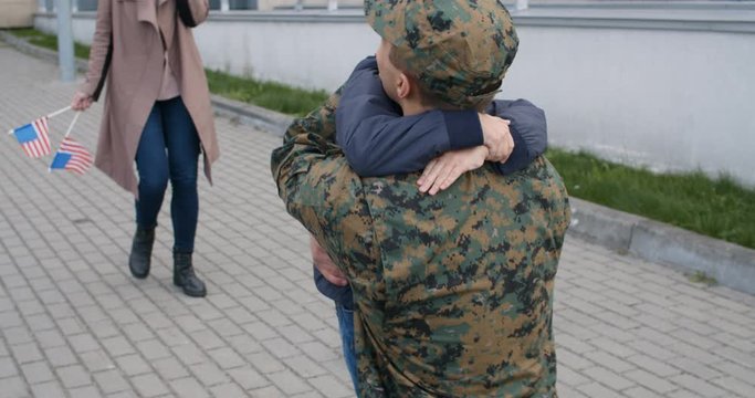 Beautiful wife and son with meeting military man outdoors. Happy little kid throwing welcome home poster and running to his military father. Family of soldier in embrace. Concept of reunion.
