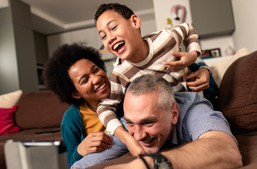 Smiling mixed race family enjoying time at home laying on sofa in living room making selfie with tablet.