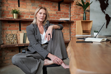 Portrait of serious confident barefoot businesswoman in loose trousers sitting at desk in own office