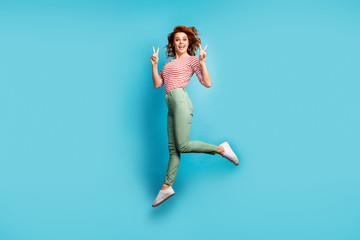 Full length profile photo of beautiful crazy lady jumping up high showing v-sign symbols rejoicing wear casual red white shirt green trousers sneakers isolated blue color background