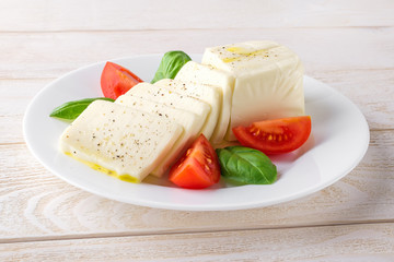 Rectangular slices of mozzarella cheese, fresh basil and quarters of tomato on a white plate over a...