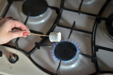 Hand holding stick with marshmallows over an open fire at gas stove in kitchen. Selective focus. 