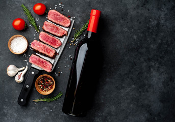  several slices of grilled steak over a meat knife with spices and a bottle of red wine on a stone background with copy space for your text