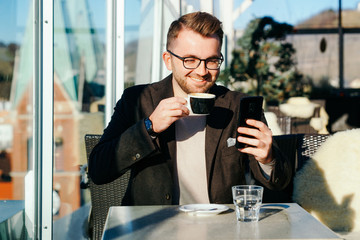 One caucasian man wearing glasses holds smartphone and drinks coffee from cup at cafe