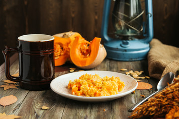  Millet porridge with pumpkin on a white plate, pumpkin slices, autumn leaves, cinnamon sticks and an antique mug with tea on an old wooden table.