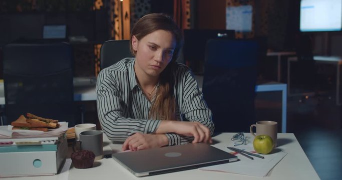 Tired female office worker closing down her laptop and banging head on arms. Stressed businesswoman looking burnout because of difficulties while working late at night in empty office