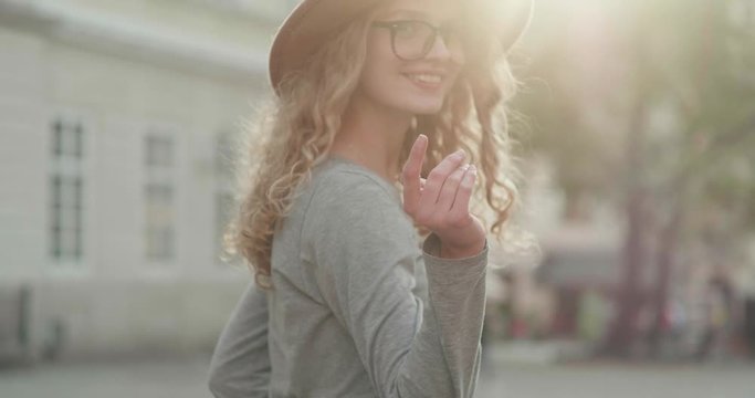 Stylish Young Woman walking in the Street while posing on Camera and calling with her Hand. Looking pretty and gorgeous. Attractive girl having beautiful curly Hair. Having Eyeglasses and trendy Hat.