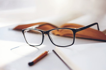 On a white table lies an open book, elegant black rimmed glasses, a notebook for notes and a pencil, illuminated by the light of the midday sun.