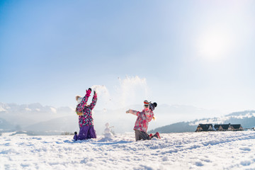 Cheerful little girls in outwear having fun and throwing snow in sunlight standing outdoors