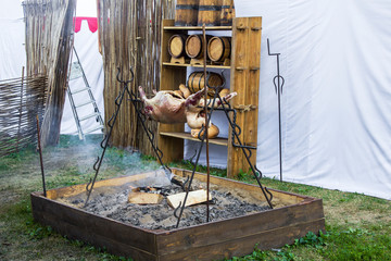 a large carcass of an animal is being cooked on a fire against the background of wooden old barrels and a tent