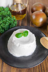 A fresh soft ricotta cheese with basil leaf on black plate, wooden tables with tomatoes, olive oil, greens. Traditional Italian food concept. Organic, homemade product.