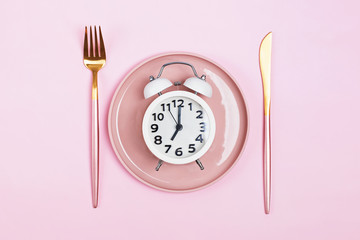 Retro alarm clock on plate with fork and knife isolated on pink background.